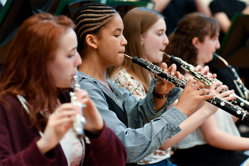 A group of young students, wearing casual clothes, performing on woodwind instruments.